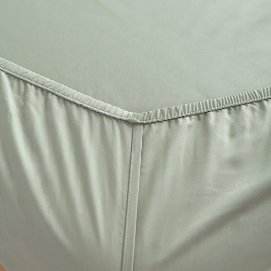 Close-up of the Sage Green bamboo fitted sheet's corner seam and elastic stitching on a wood bed frame.